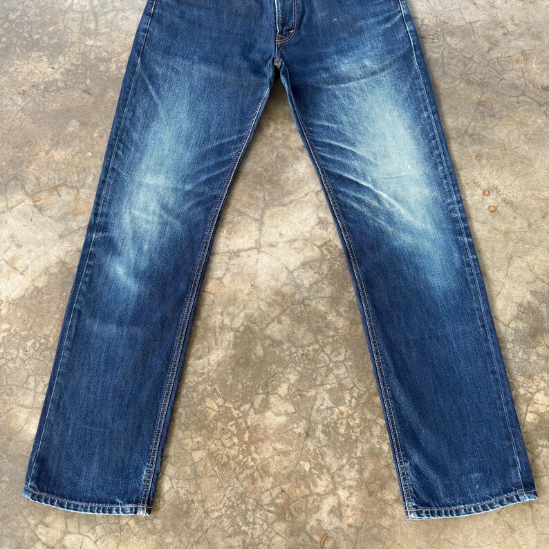 May include: A pair of blue denim jeans with a faded wash effect. The jeans have a classic five-pocket design and a straight leg cut. The denim has a worn appearance with lighter areas on the thighs and knees, suggesting wear and tear.
