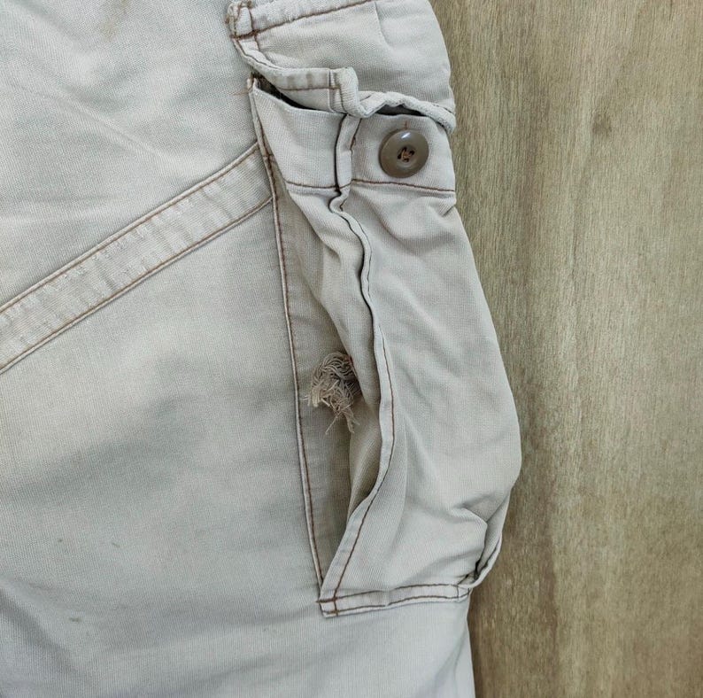 May include: Close-up of tan cargo pants with a visible pocket and button detail. The pocket has a distressed area, showing wear and tear. The pants have visible stitching and a neutral color.