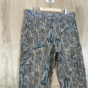 May include: Camouflage cargo trousers with a button and zip fastening, featuring multiple pockets. The trousers have a pattern of brown, green, and blue tree branches and leaves. The trousers are hanging on a wooden hanger.