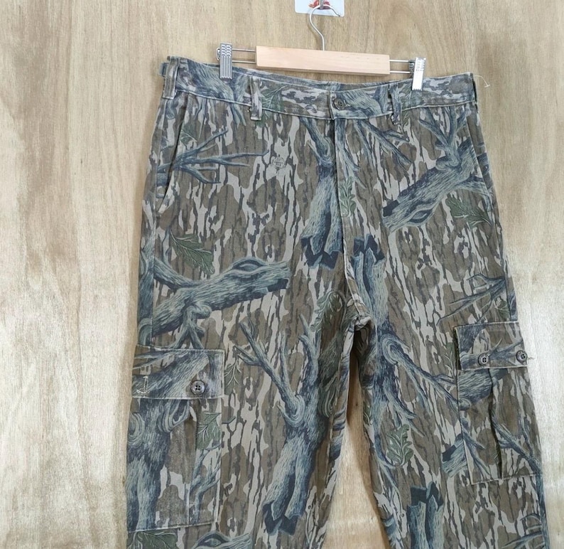 May include: Camouflage cargo trousers with a button and zip fastening, featuring multiple pockets. The trousers have a pattern of brown, green, and blue tree branches and leaves. The trousers are hanging on a wooden hanger.