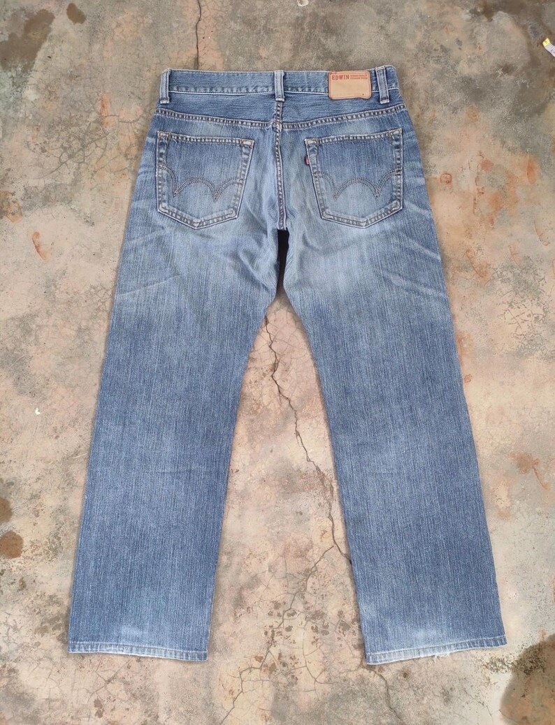 May include: Light blue denim jeans with a faded wash. The jeans feature classic five-pocket styling, belt loops, and a leather patch on the back waistband. The back pockets have a distinctive curved design. The jeans are laid flat.