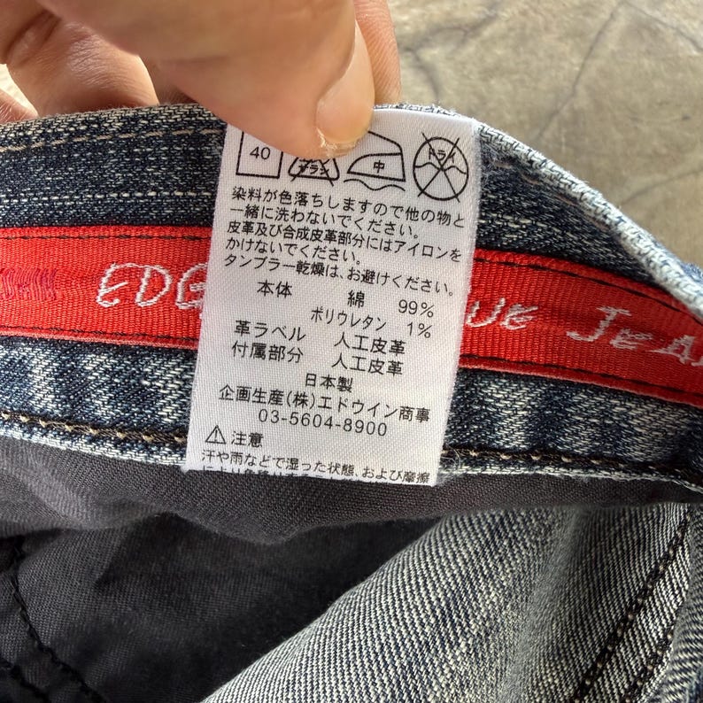 以下が含まれることがあります： デニム生地のクローズアップ。赤いラベルと白いタグが付いています。タグには日本語のテキストとケアマークがあります。デニムは青色で、色あせた外観です。