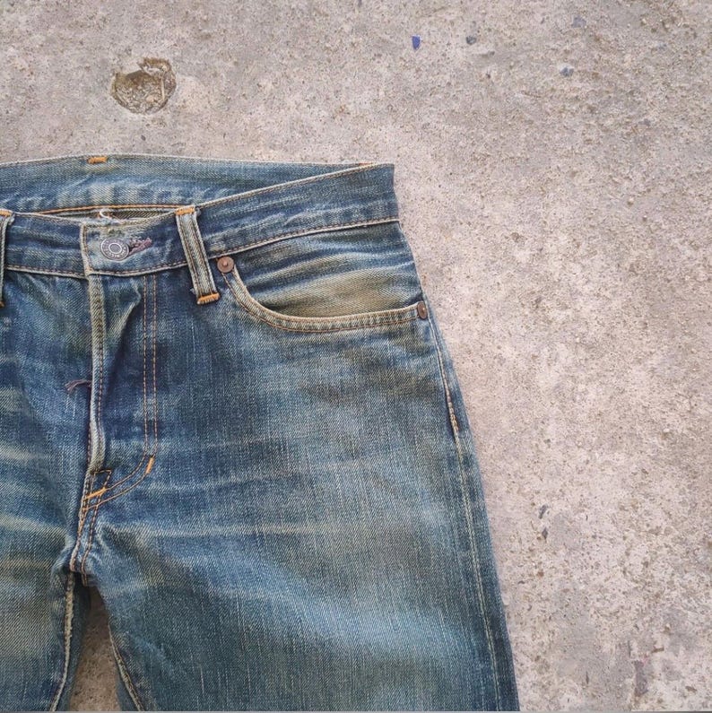 May include: Close-up of vintage blue denim jeans. The jeans show signs of wear with faded areas and subtle color variations. Features include a button closure, front pockets, and visible stitching. The overall aesthetic is a classic, worn look.