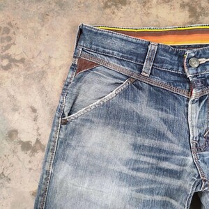 以下が含まれることがあります： 色あせたウォッシュ加工のブルーデニムジーンズのクローズアップ。ポケットのブラウンレザーアクセント、ボタン留め、ストライプのウエストバンドが特徴です。ジーンズはクラシックな5ポケットデザインです。