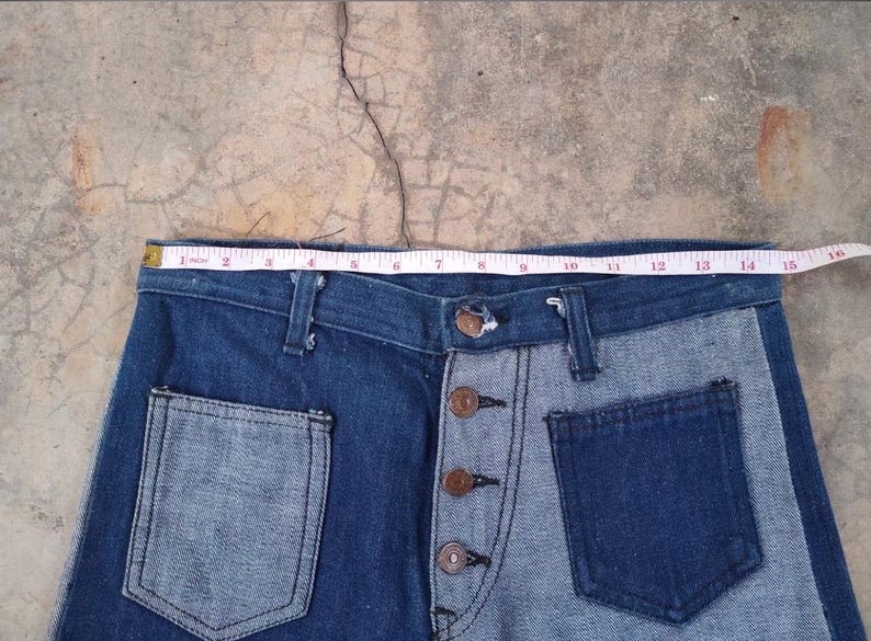 May include: Two-tone denim jeans with a button fly and front pockets. The jeans feature a light blue and dark blue color scheme. A measuring tape shows the waist is approximately 15 inches wide.