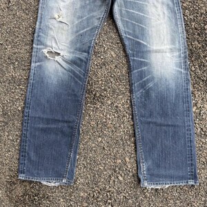 May include: Faded blue denim jeans with distressed details, including a large hole on the left leg. The jeans have a classic five-pocket design and a straight leg cut. The denim shows wear and tear.