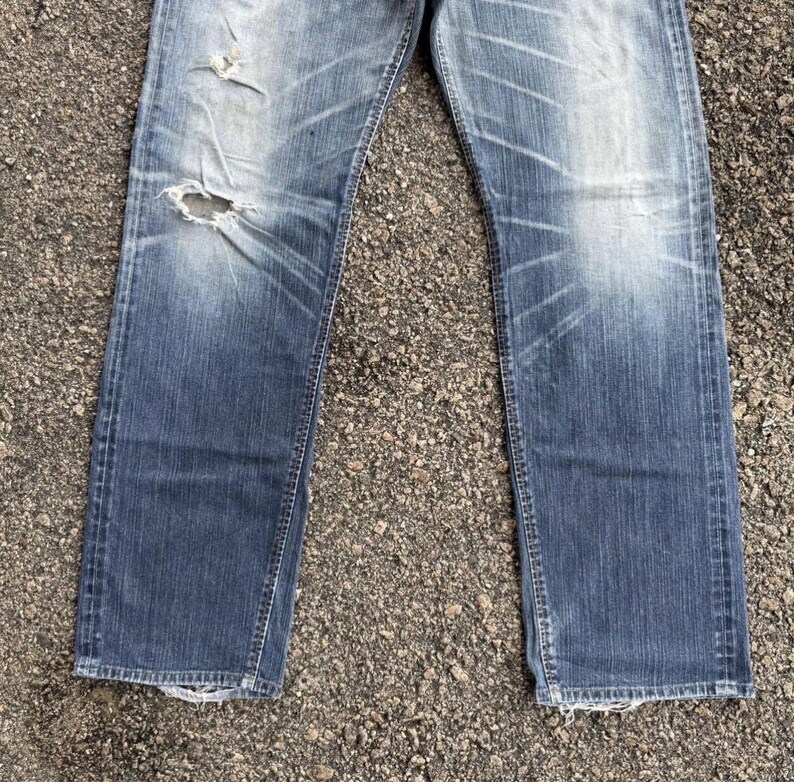 May include: Faded blue denim jeans with distressed details, including a large hole on the left leg. The jeans have a classic five-pocket design and a straight leg cut. The denim shows wear and tear.