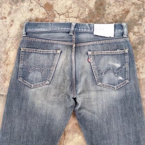 Puede incluir: Vaqueros de mezclilla azul descolorido con dise&ntilde;o cl&aacute;sico de cinco bolsillos. Incluye costuras blancas en los bolsillos traseros, una etiqueta roja y un parche de cuero. Los vaqueros tienen un aspecto desgastado y vintage.