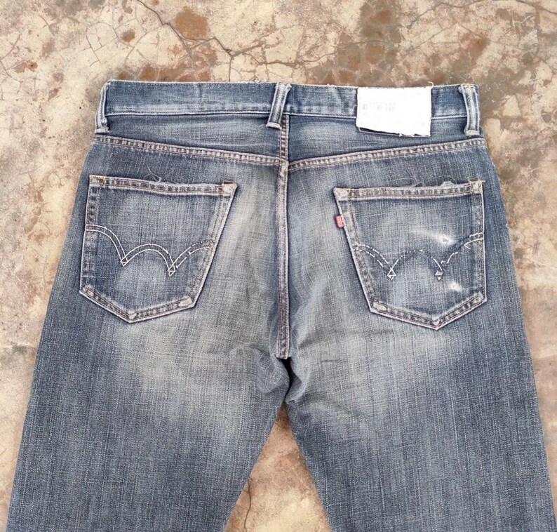 Puede incluir: Vaqueros de mezclilla azul descolorido con dise&ntilde;o cl&aacute;sico de cinco bolsillos. Incluye costuras blancas en los bolsillos traseros, una etiqueta roja y un parche de cuero. Los vaqueros tienen un aspecto desgastado y vintage.