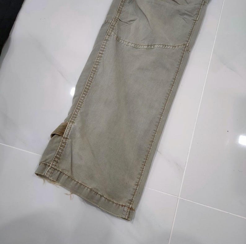 May include: Close-up of olive green cargo pants. The pants have a faded appearance with visible stitching and a small pocket detail. The fabric appears to be a durable material, suitable for outdoor activities. The pants are slightly distressed at the hem.