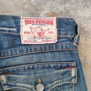 May include: Close-up of blue denim jeans with a True Religion brand label. The label has red text and a graphic of a figure playing a guitar. The jeans feature a button closure and back pocket detail.