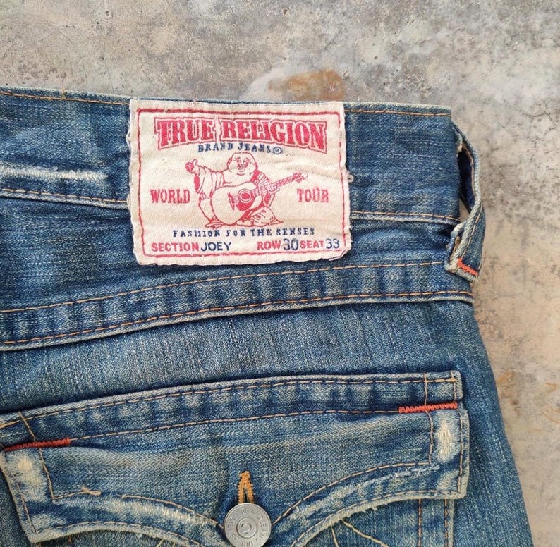 May include: Close-up of blue denim jeans with a True Religion brand label. The label has red text and a graphic of a figure playing a guitar. The jeans feature a button closure and back pocket detail.