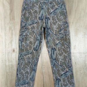 May include: Camouflage cargo trousers in shades of green, brown, and grey, with a tree bark pattern. The trousers have multiple pockets and a drawstring at the bottom of the legs. The trousers are hanging on a wooden hanger.