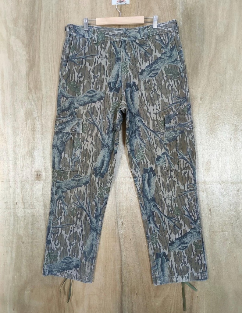 May include: Camouflage cargo trousers in shades of green, brown, and grey, with a tree bark pattern. The trousers have multiple pockets and a drawstring at the bottom of the legs. The trousers are hanging on a wooden hanger.