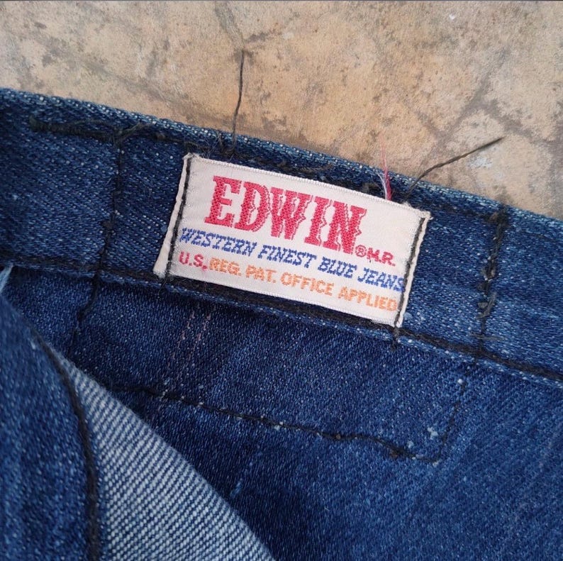 May include: Close-up of a section of dark blue denim jeans. A rectangular tag with the brand name "EDWIN" in red and white is sewn onto the jeans. The tag also includes the text "WESTERN FINEST BLUE JEANS" and "U.S. REG. PAT. OFFICE APPLIED."