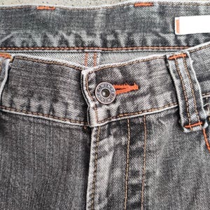 K&ouml;nnte beinhalten: Nahaufnahme einer grauen Jeans. Die Jeans hat orangefarbene N&auml;hte und einen Knopf mit dem Markennamen "Edwin". Der Knopf ist &uuml;ber dem Hosenschlitz zentriert. Die Jeans hat ein klassisches F&uuml;nf-Taschen-Design.