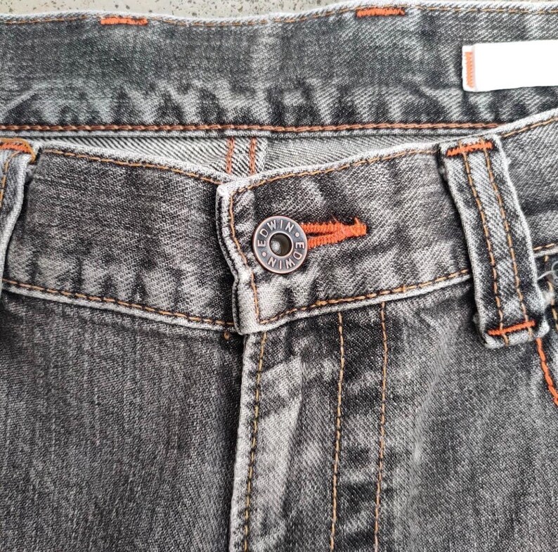 K&ouml;nnte beinhalten: Nahaufnahme einer grauen Jeans. Die Jeans hat orangefarbene N&auml;hte und einen Knopf mit dem Markennamen "Edwin". Der Knopf ist &uuml;ber dem Hosenschlitz zentriert. Die Jeans hat ein klassisches F&uuml;nf-Taschen-Design.
