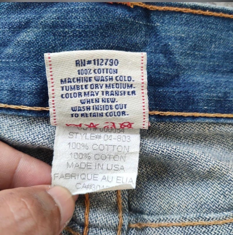 May include: Close-up of a denim garment with a tag. The tag reads "RN#112790" and includes washing instructions: "MACHINE WASH COLO. TUMBLE DRY MEDIUM." Additional text includes "100% COTTON" and "MADE IN USA."