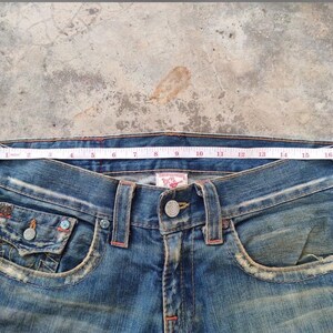 May include: Close-up of blue denim jeans with a measuring tape across the waist. The jeans have a button closure, front pockets, and decorative stitching. The waist measures approximately 40 cm across.