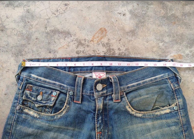 May include: Close-up of blue denim jeans with a measuring tape across the waist. The jeans have a button closure, front pockets, and decorative stitching. The waist measures approximately 40 cm across.