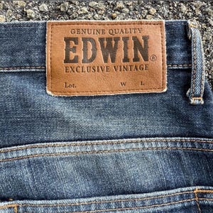 Pu&ograve; includere: Primo piano di un paio di jeans blu. Una toppa in pelle marrone &egrave; cucita sul retro dei jeans, con il marchio "EDWIN" e il testo "GENUINE QUALITY" e "EXCLUSIVE VINTAGE" stampati sopra. I jeans hanno un classico design a cinque tasche.