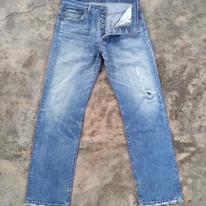 May include: Light blue denim jeans with a button fly and distressed details. The jeans have a faded wash, a ripped knee, and raw, unfinished hems. The classic five-pocket design is visible.