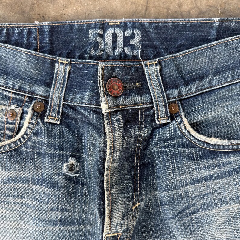 May include: Close-up of blue denim jeans with the number "503" printed on the waistband. The jeans have a button closure and rivets. The denim is faded and distressed, showing signs of wear.