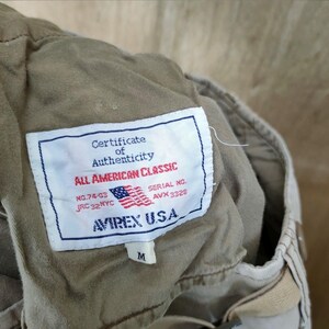 May include: Close-up of a tan garment tag with the text "Certificate of Authenticity" and "ALL AMERICAN CLASSIC". The tag includes the American flag and the brand "AVIREX U.S.A.". The tag also has the size "M".