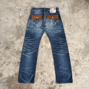 Op de afbeelding: Blauwe denim jeans met bruine, su&egrave;de-achtige achterzakken en een rood Levi's-label. De jeans hebben een vervaagde, distressed look met witte stiksels en een knoopsluiting. De achterzakken hebben een knoopsluiting.