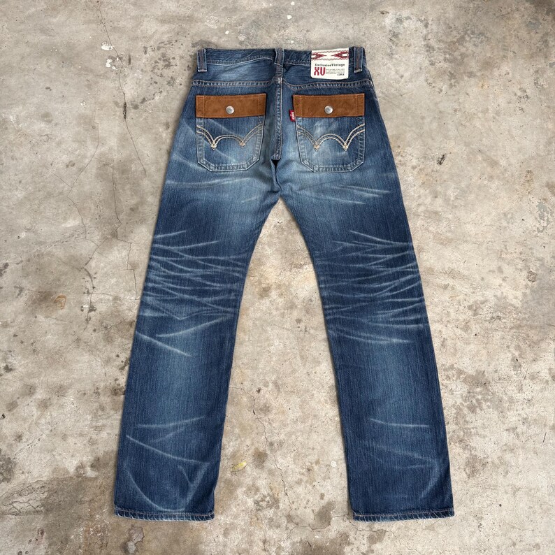 Op de afbeelding: Blauwe denim jeans met bruine, su&egrave;de-achtige achterzakken en een rood Levi's-label. De jeans hebben een vervaagde, distressed look met witte stiksels en een knoopsluiting. De achterzakken hebben een knoopsluiting.