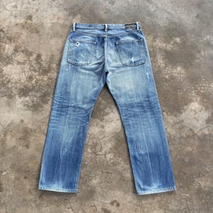 Pu&ograve; includere: Jeans in denim blu chiaro con un aspetto usurato e vissuto. I jeans hanno un classico design a cinque tasche, con segni di usura visibili, tra cui un piccolo foro sulla gamba sinistra. I jeans sono stesi piatti, mostrando le tasche posteriori e i passanti per la cintura.