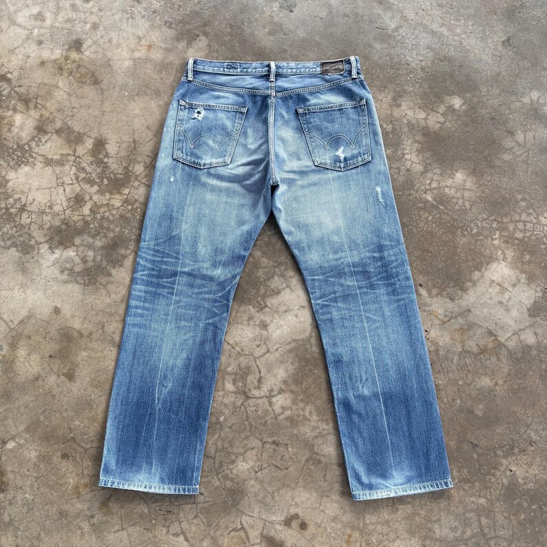 Pu&ograve; includere: Jeans in denim blu chiaro con un aspetto usurato e vissuto. I jeans hanno un classico design a cinque tasche, con segni di usura visibili, tra cui un piccolo foro sulla gamba sinistra. I jeans sono stesi piatti, mostrando le tasche posteriori e i passanti per la cintura.