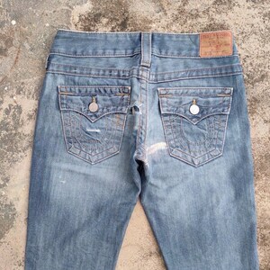 May include: Faded light blue denim jeans. They have back pockets with decorative stitching, silver button closures, and a brown leather brand patch. The jeans have a classic five-pocket design and a worn appearance.