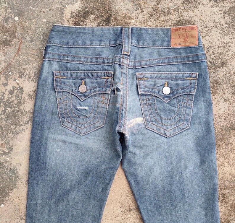 May include: Faded light blue denim jeans. They have back pockets with decorative stitching, silver button closures, and a brown leather brand patch. The jeans have a classic five-pocket design and a worn appearance.