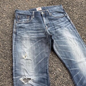 May include: Faded blue denim jeans with distressed details, including a ripped knee. The jeans have a classic five-pocket design and a button and zipper closure. The brand name "Edwin" is visible on a small tag.