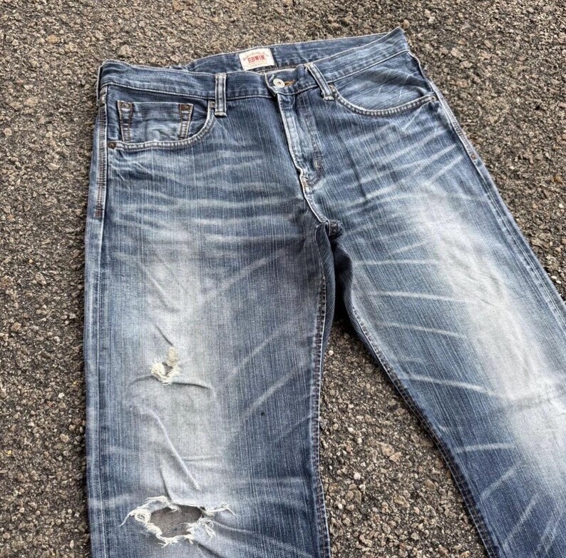 May include: Faded blue denim jeans with distressed details, including a ripped knee. The jeans have a classic five-pocket design and a button and zipper closure. The brand name "Edwin" is visible on a small tag.