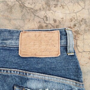 May include: Close-up of blue denim jeans with a brown leather patch. The patch is rectangular and has a worn appearance. The jeans have yellow stitching and a classic design. The background is a textured, light-colored surface.