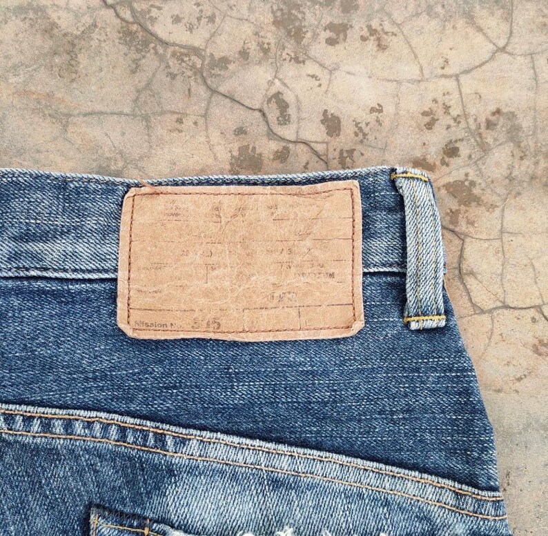 May include: Close-up of blue denim jeans with a brown leather patch. The patch is rectangular and has a worn appearance. The jeans have yellow stitching and a classic design. The background is a textured, light-colored surface.