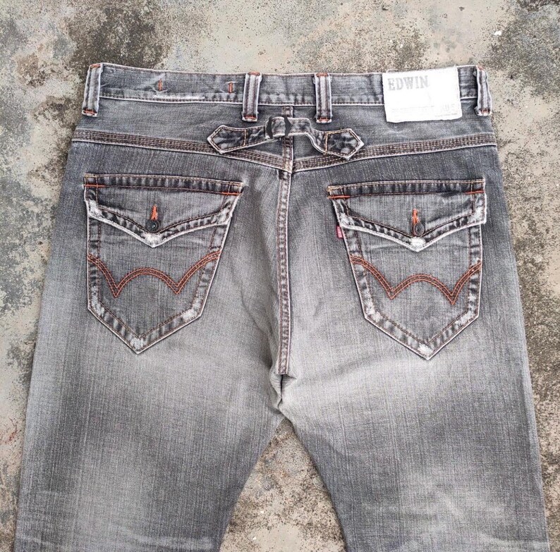 K&ouml;nnte beinhalten: Verblasste graue Jeans mit einem einzigartigen Design der Ges&auml;&szlig;taschen und roten N&auml;hten. Die Jeans haben ein markantes R&uuml;ckenteil und den Markennamen "EDWIN" auf einem Etikett. Die Taschen haben eine stilisierte "W"-Form.