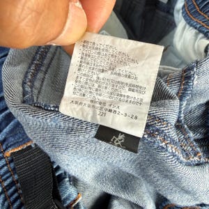 May include: Close-up of a denim garment with a white tag featuring Japanese text and care instructions. The tag also includes the address of a company in Osaka, Japan, and the number 221. The garment is blue.