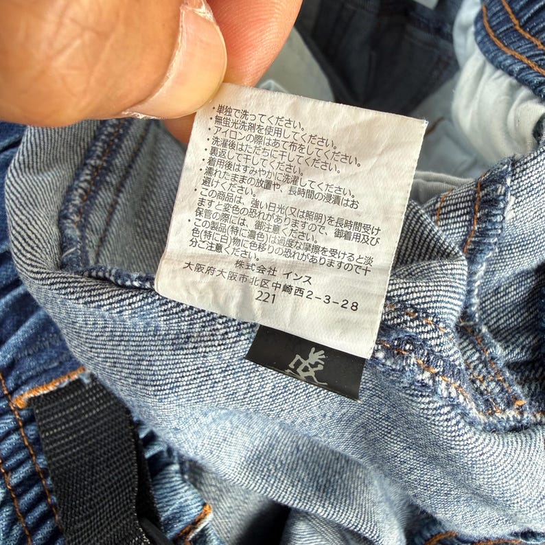 May include: Close-up of a denim garment with a white tag featuring Japanese text and care instructions. The tag also includes the address of a company in Osaka, Japan, and the number 221. The garment is blue.