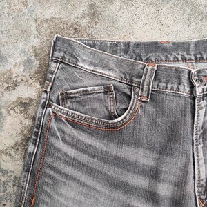K&ouml;nnte beinhalten: Nahaufnahme einer grauen Jeans. Die Jeans hat ein klassisches F&uuml;nf-Taschen-Design mit orangefarbenen N&auml;hten. Der Stoff weist ein abgenutztes, strukturiertes Aussehen auf, und die Jeans ist am Bund gekn&ouml;pft.