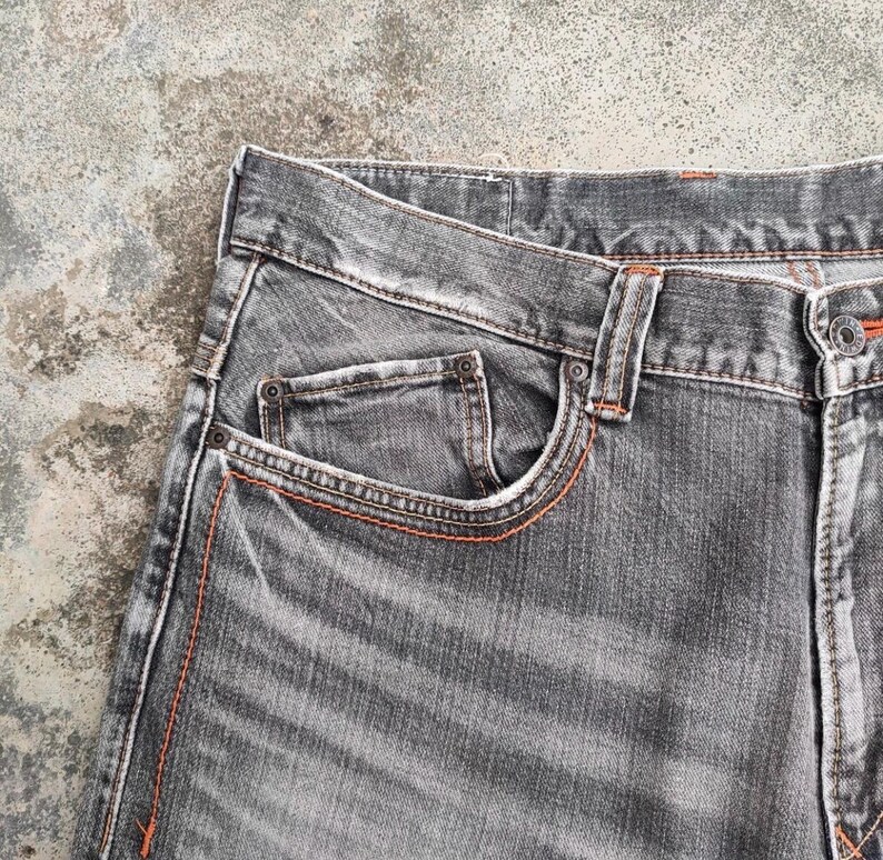 K&ouml;nnte beinhalten: Nahaufnahme einer grauen Jeans. Die Jeans hat ein klassisches F&uuml;nf-Taschen-Design mit orangefarbenen N&auml;hten. Der Stoff weist ein abgenutztes, strukturiertes Aussehen auf, und die Jeans ist am Bund gekn&ouml;pft.