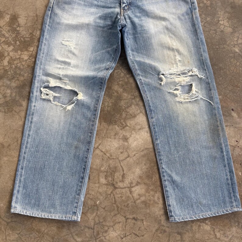 May include: Light blue denim jeans with distressed details, including large rips at the knees. The jeans have a faded wash and a classic five-pocket design. The fabric shows signs of wear.