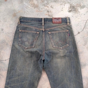 May include: Faded blue denim jeans with a classic five-pocket design. The jeans feature a brown leather patch on the back and a distinctive stitched design on the back pockets. The denim has a worn, vintage look.