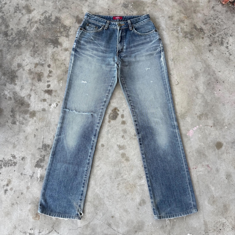 May include: Faded blue denim jeans with a distressed look. The jeans have a classic five-pocket design, a button and zipper closure, and a small rip on the left knee. The jeans are slightly flared at the bottom.