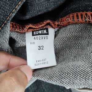 Pu&ograve; includere: Primo piano di un capo in denim con cucitura arancione. Un'etichetta bianca mostra il marchio "EDWIN", la taglia "32" e il modello "402XVS". Sono visibili anche altri codici prodotto.