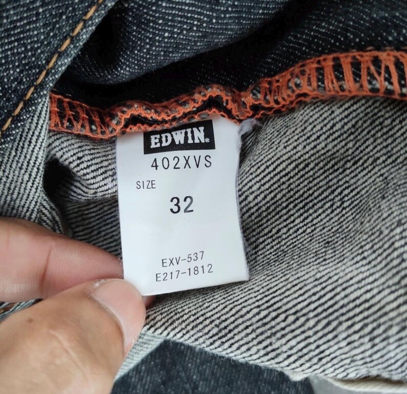 Pu&ograve; includere: Primo piano di un capo in denim con cucitura arancione. Un'etichetta bianca mostra il marchio "EDWIN", la taglia "32" e il modello "402XVS". Sono visibili anche altri codici prodotto.
