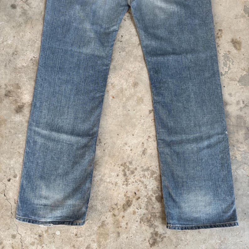 May include: Light blue denim jeans with a classic five-pocket design. The jeans have a straight leg cut and show signs of wear and fading, giving them a vintage look. The fabric has a textured appearance.