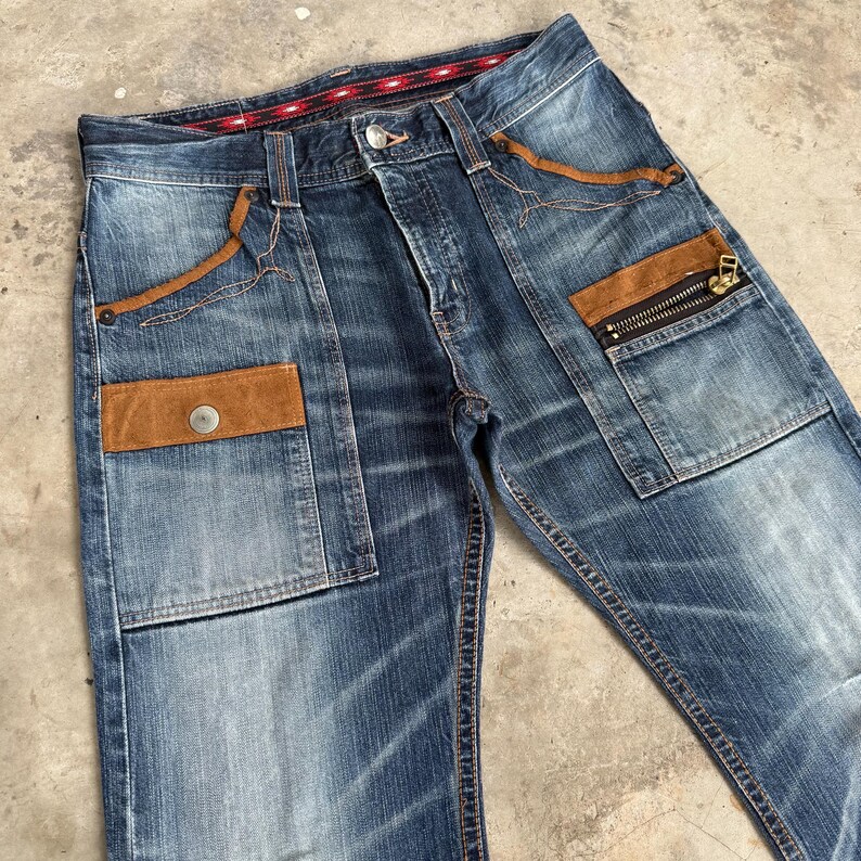 Op de afbeelding: Blauwe spijkerbroek met bruine su&egrave;de zakaccenten en een zak met ritssluiting. De jeans heeft een knoopsluiting en een rood-zwarte patroon tailleband detail. De jeans is licht vervaagd en heeft een versleten uiterlijk.